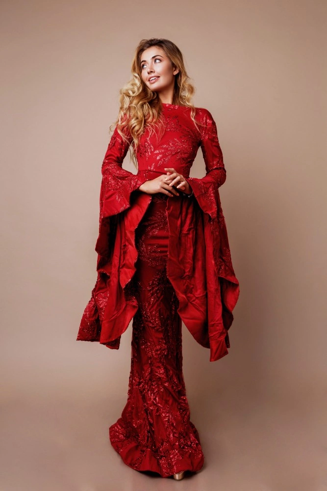 “A woman posing in a studio against a neutral beige background, wearing an elegant deep red, full-length gown with intricate textured detailing. The dress features long sleeves with dramatic draped fabric and a fitted silhouette. Her long, wavy hair falls over one shoulder as she looks slightly upward, creating a refined and graceful fashion portrait.