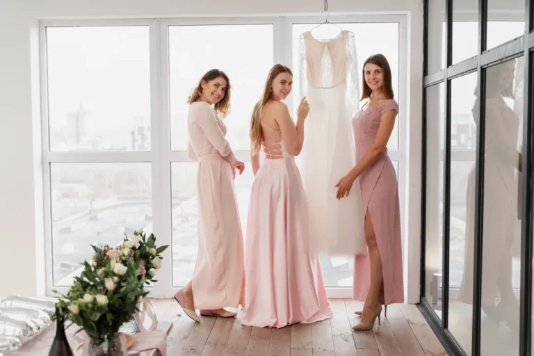 Three women wearing elegant pink prom dresses standing indoors near a window, showcasing soft pastel prom dress styles and feminine evening fashion.