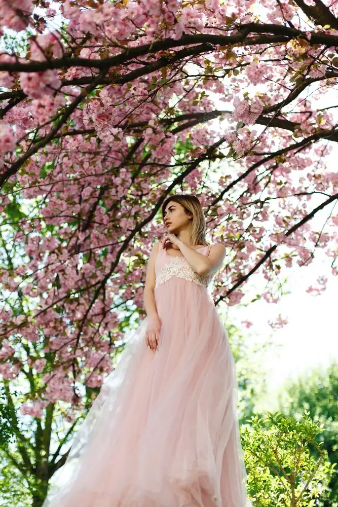 Woman wearing a soft pink tulle prom dress standing beneath blooming cherry blossom trees, showcasing a romantic and dreamy evening dress style.
