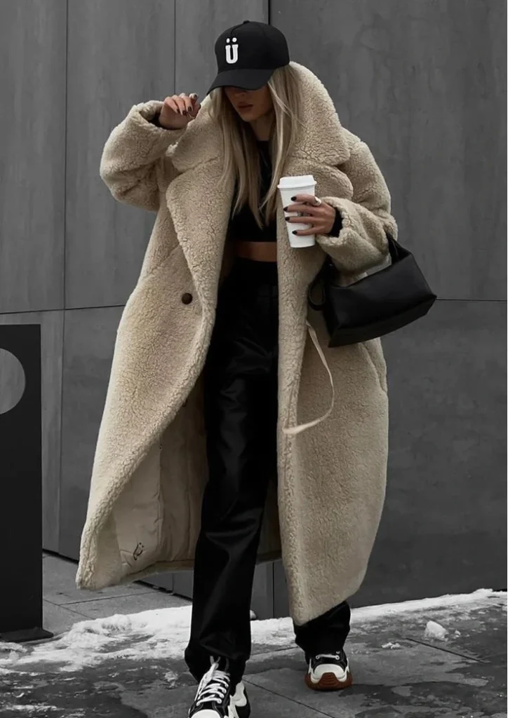 Street-style winter outfit featuring a long teddy coat, black crop top and pants, baseball cap, sneakers, and a tote bag for a trendy cold-weather look.