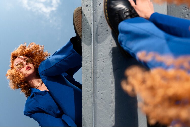 Fashion-forward woman with curly hair wearing a bold blue suit and sunglasses, posing creatively against a concrete structure under a clear sky.
