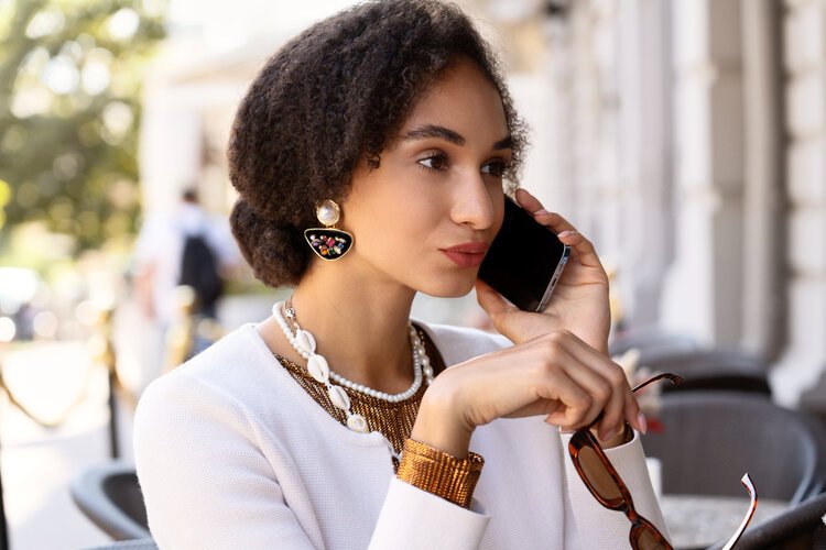Woman wearing elegant demi-fine jewellery, including statement earrings and layered necklaces, while talking on her phone outdoors.