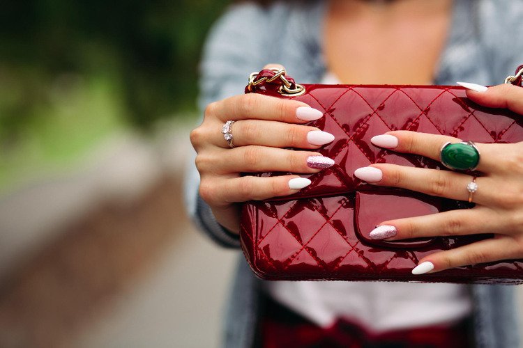 Close-up of manicured hands holding a red quilted handbag, featuring almond-shaped white nails with a glitter accent nail and statement rings.
