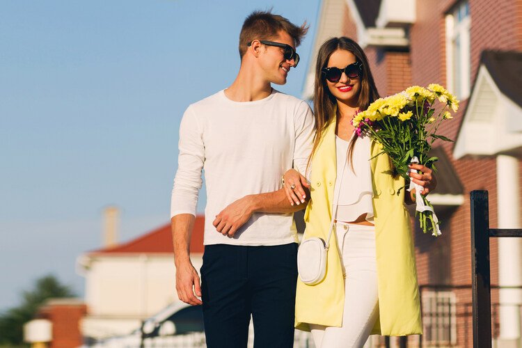 A stylish couple walking outdoors on a sunny day, wearing coordinated casual outfits—one in a white long-sleeve top and dark trousers, the other in a yellow coat and white outfit—smiling and holding a bouquet of flowers, creating a relaxed first-date vibe.