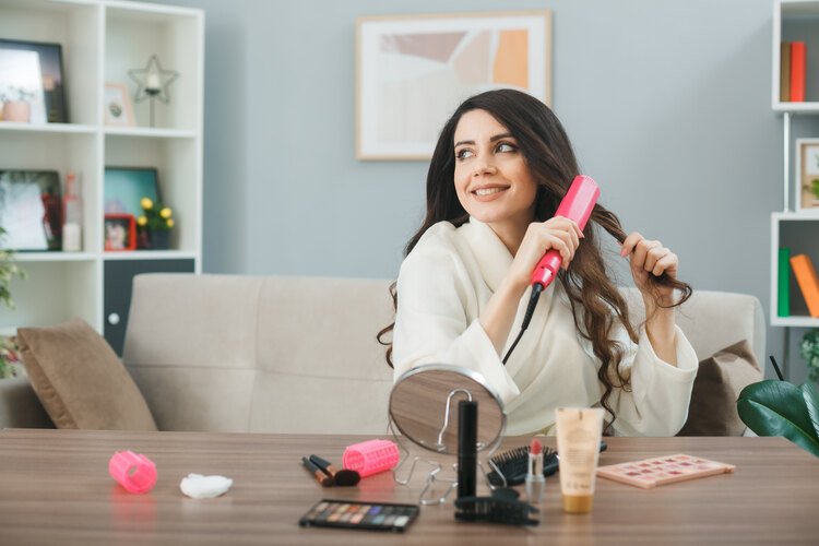 Woman using a pink hair waver at home, creating loose waves while sitting at a dressing table with makeup and styling tools.